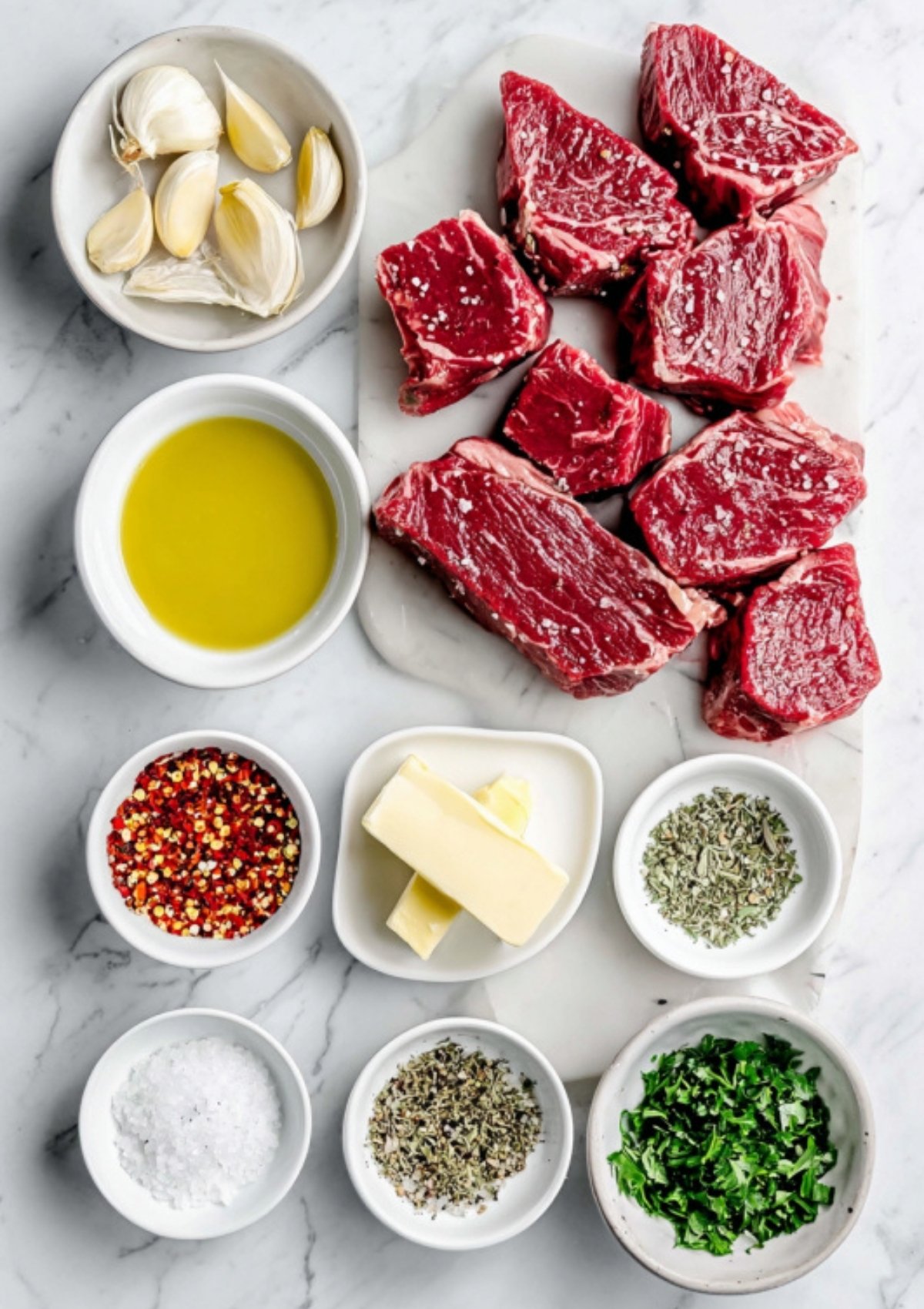 Overhead view of ingredients for garlic butter steak bites including cubed steak, garlic cloves, olive oil, butter, red pepper flakes, herbs, and fresh parsley arranged neatly on a marble surface.