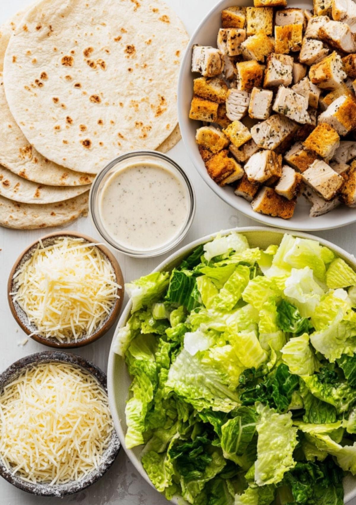 Overhead view of tortillas, romaine lettuce, grilled chicken cubes, Parmesan cheese, and Caesar dressing arranged on a table.