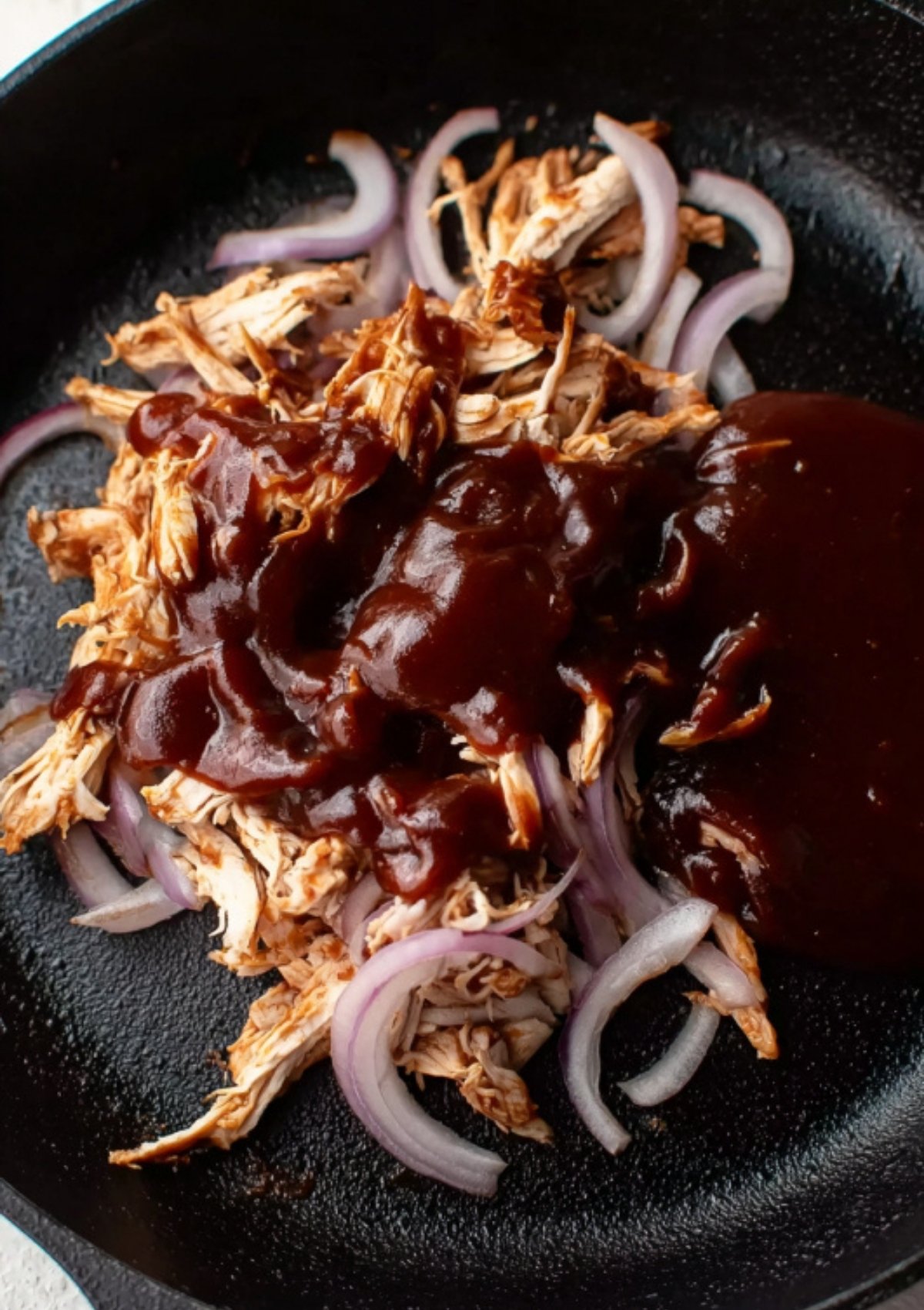 Shredded chicken and sliced red onions mixed with rich BBQ sauce in a black skillet, ready to cook.