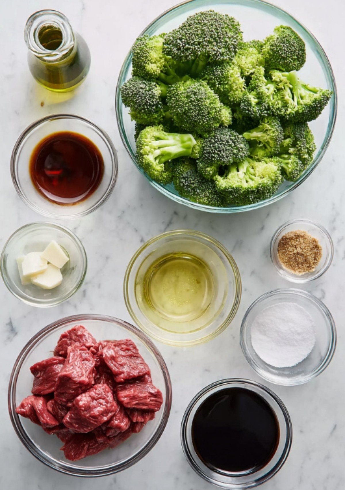 Overhead shot of bowls with beef, broccoli, soy sauce, and stir-fry ingredients on a marble counter.