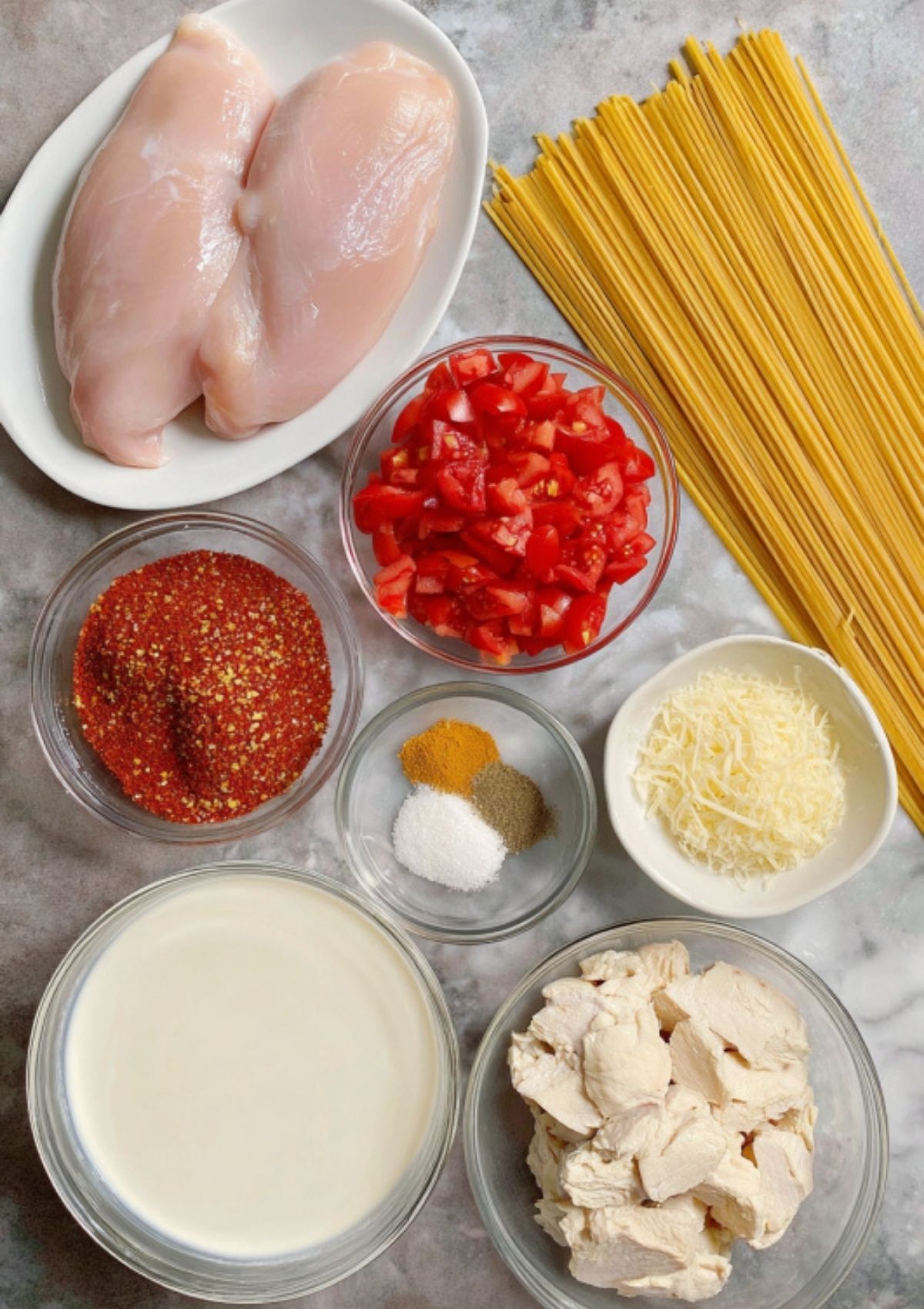 Overhead shot of ingredients for Cajun chicken Alfredo including chicken breasts, pasta, spices, cream, tomatoes, and cheese.