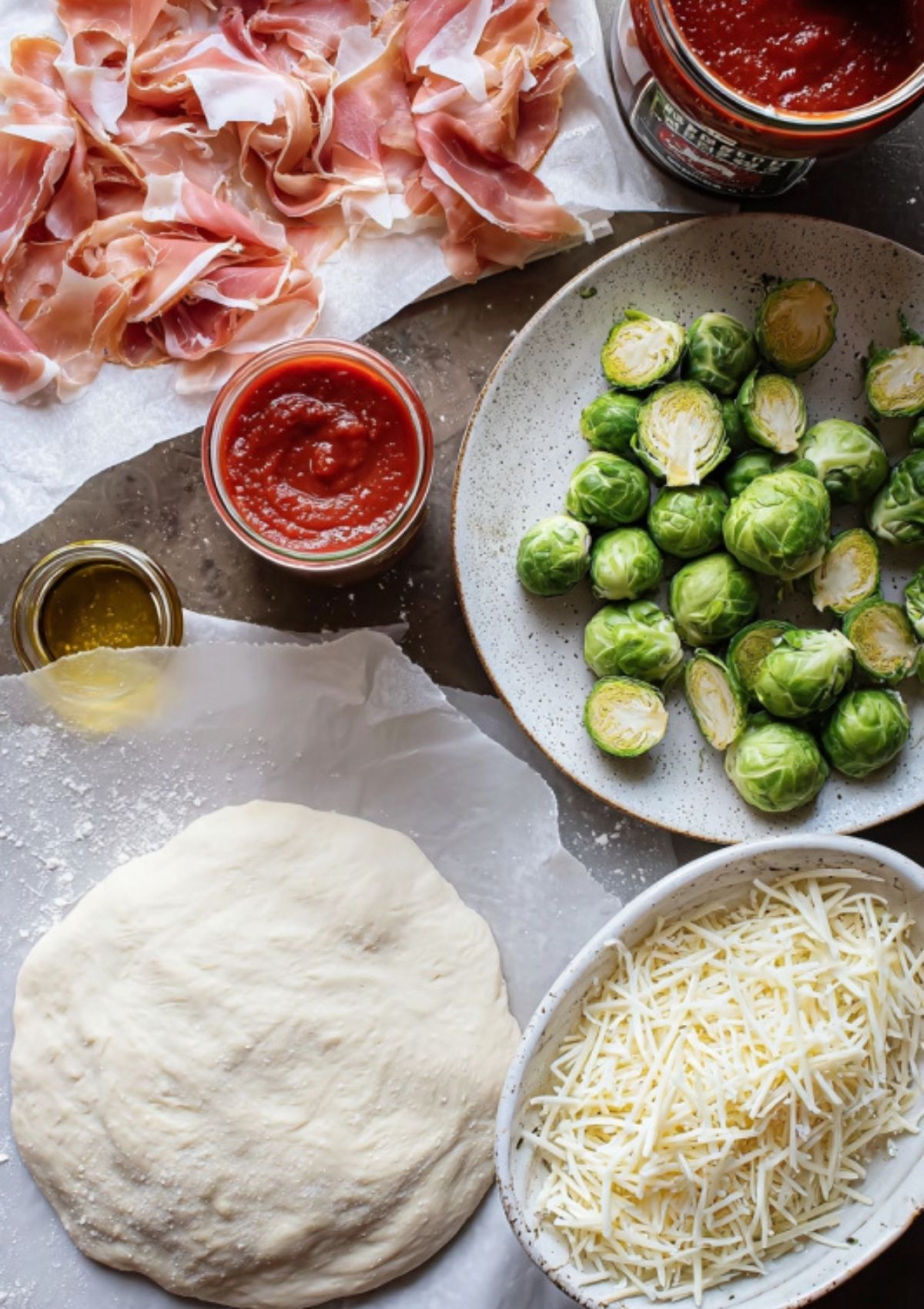 Overhead view of pizza dough, shredded cheese, prosciutto, Brussels sprouts, tomato sauce, and olive oil ready for pizza prep.