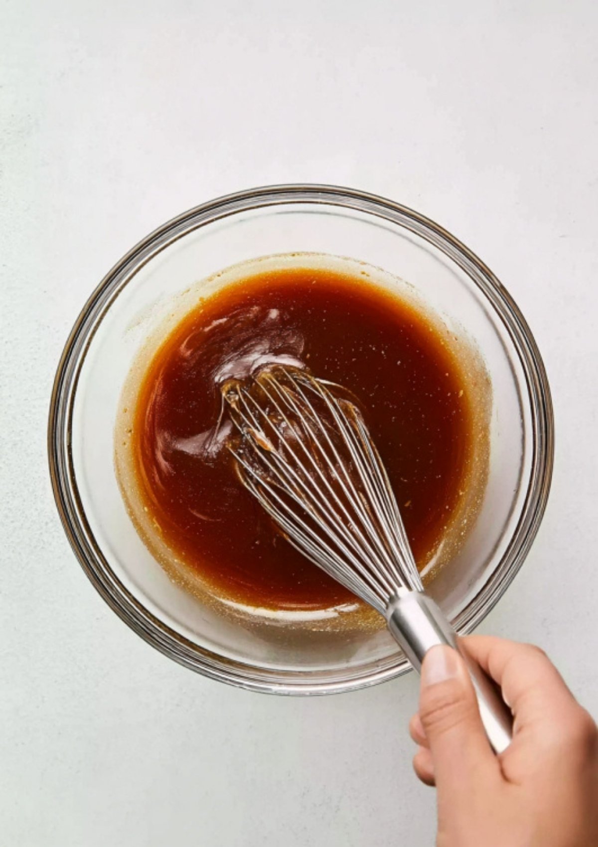 Hand whisking a glossy brown sauce in a glass bowl for beef and broccoli stir-fry.