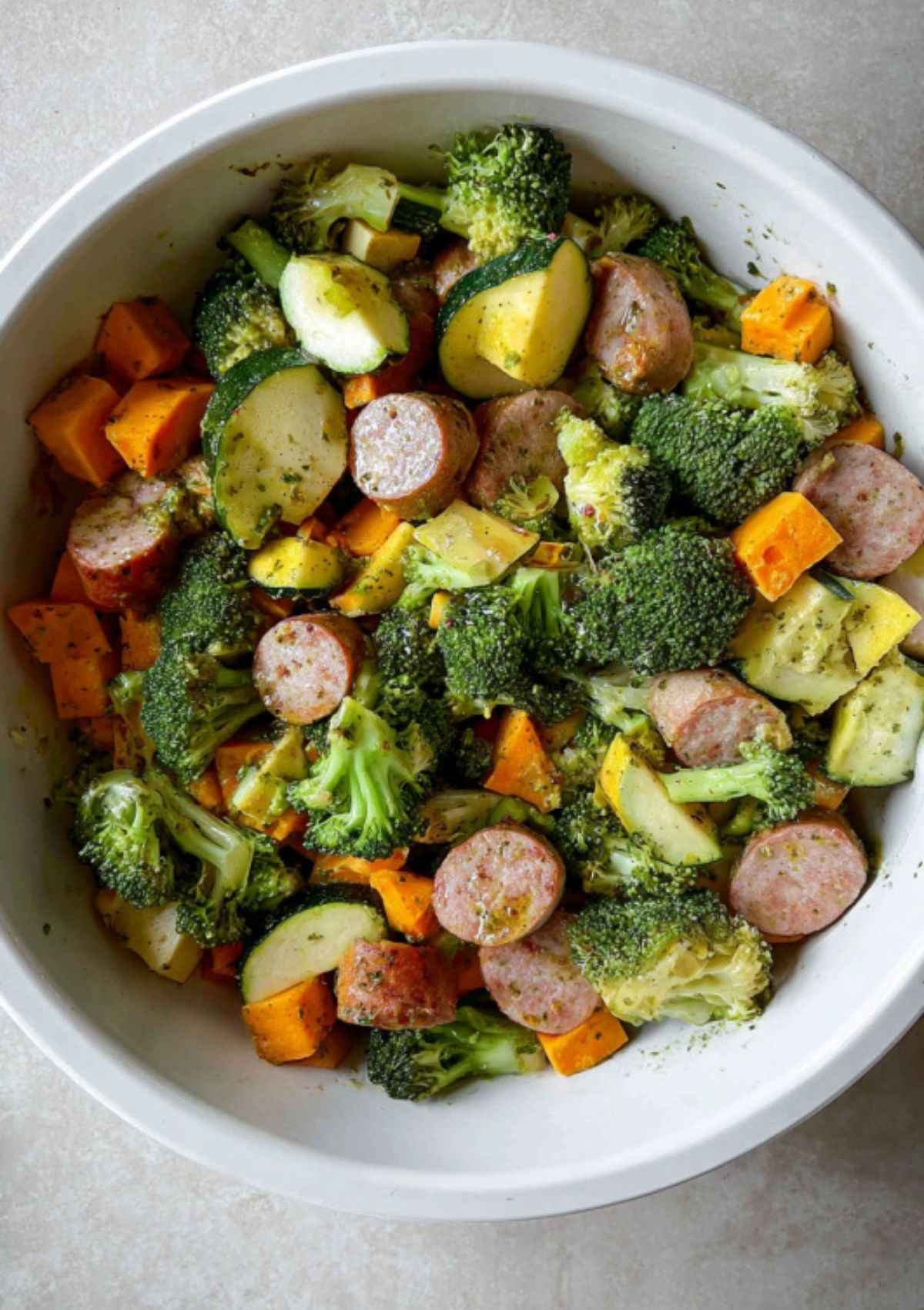 Bowl of sausage slices, broccoli, zucchini, and sweet potato tossed in olive oil and herbs, ready for roasting.