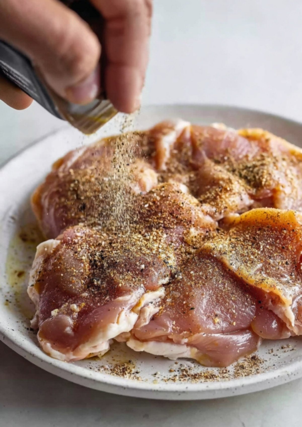 Close-up of raw chicken thighs being seasoned with salt, pepper, and spices on a white plate.
