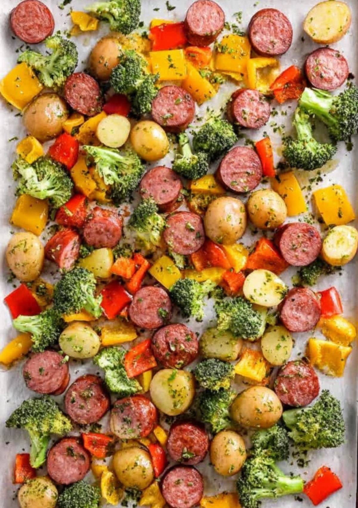 Uncooked sausage, broccoli, bell peppers, and potatoes spread evenly on a baking sheet with herbs and oil.