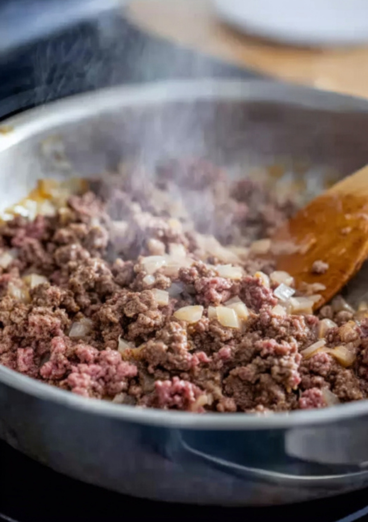 Ground beef sizzling with chopped onions in a hot skillet, stirred with a wooden spoon for slider filling.