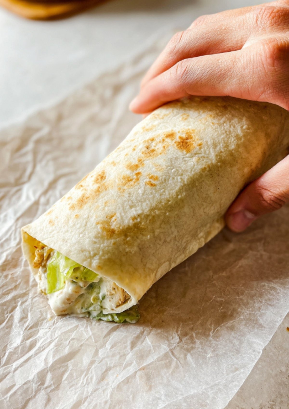 Hand rolling a flour tortilla filled with chicken Caesar salad mixture on parchment paper.