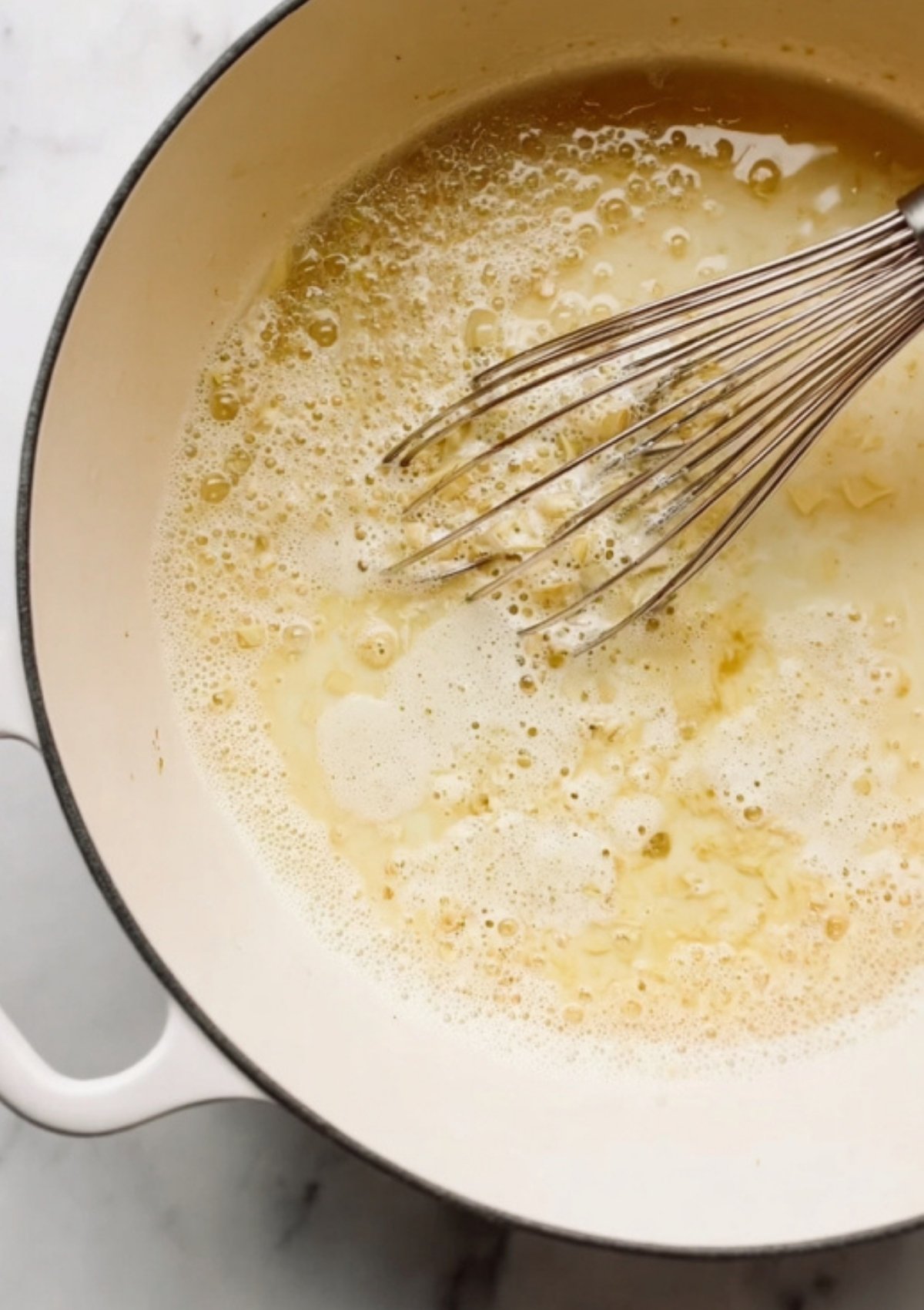 White Dutch oven with melted butter and garlic being whisked as the first step for a homemade cream sauce.