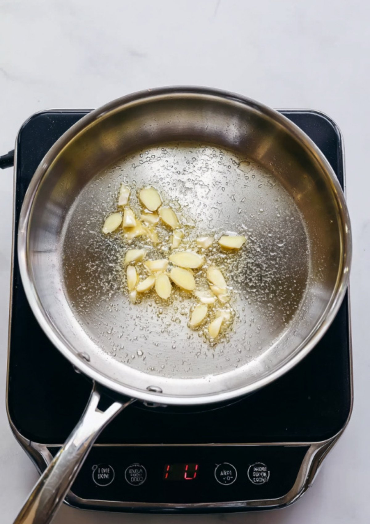 Sliced garlic sautéing in oil in a stainless-steel skillet on a stovetop.