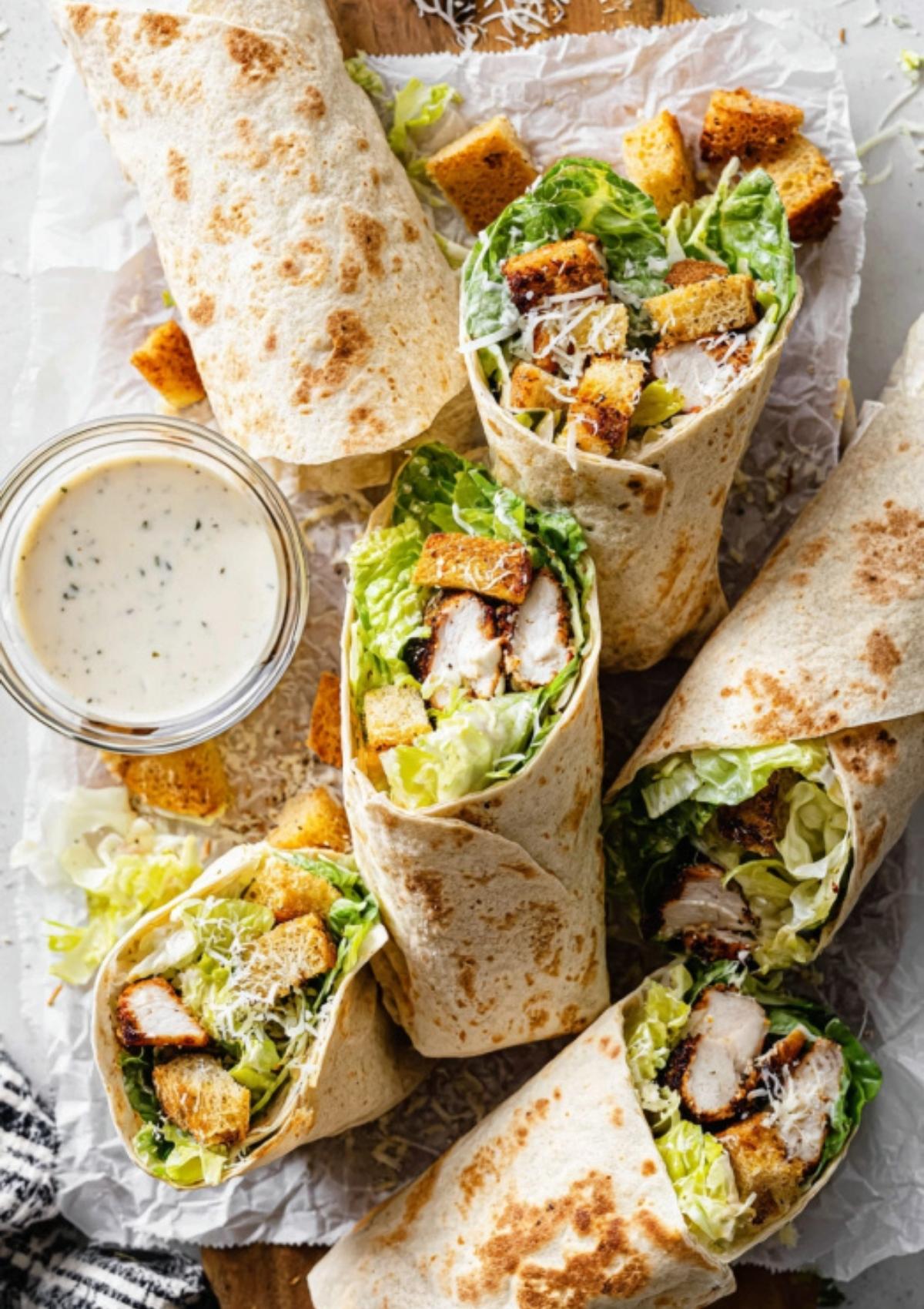 Platter of freshly made chicken Caesar wraps served with a side of creamy dressing and sprinkled Parmesan cheese.
