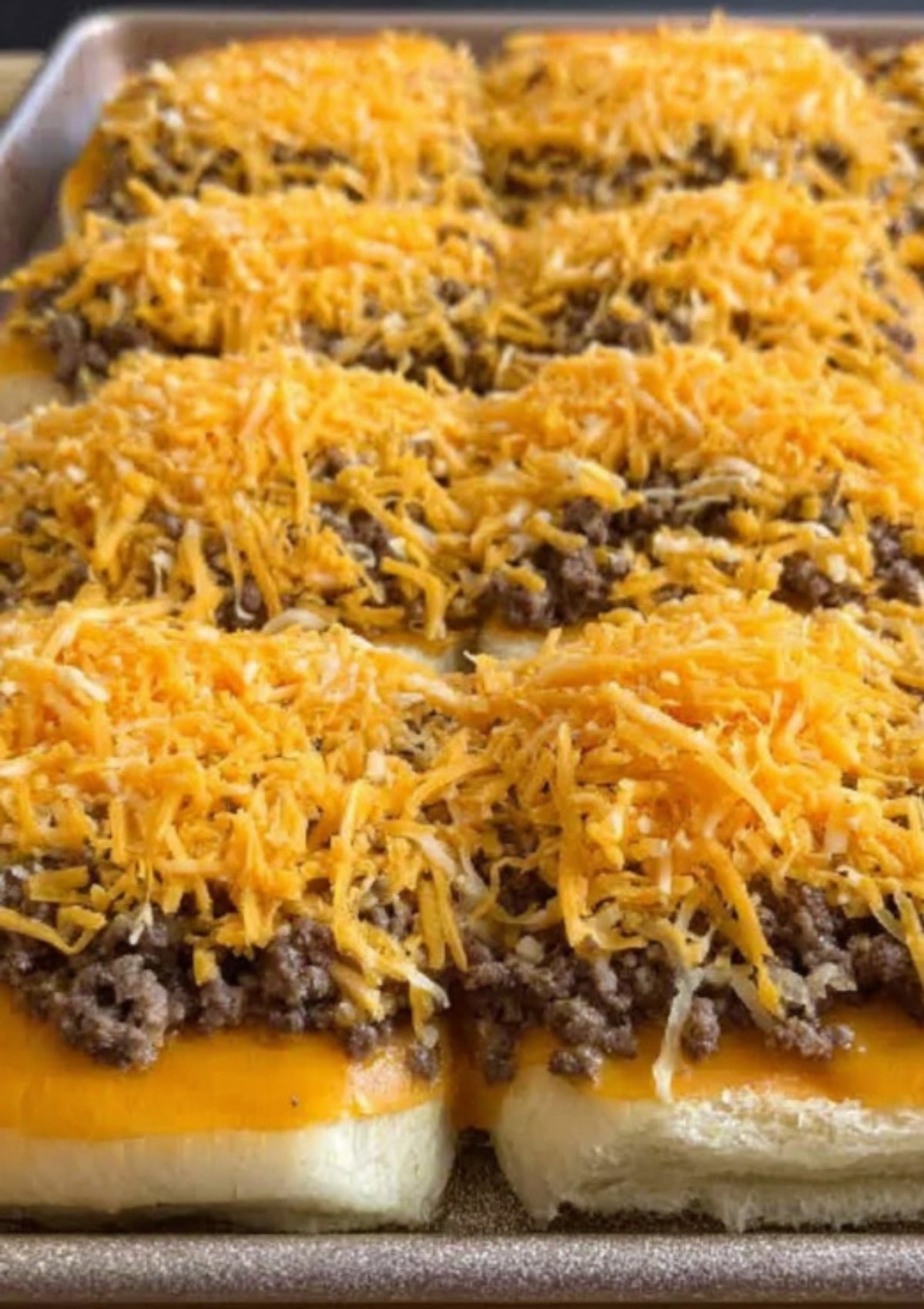 Slider buns topped with cooked ground beef and a generous layer of shredded cheddar cheese before baking for cheeseburger sliders.