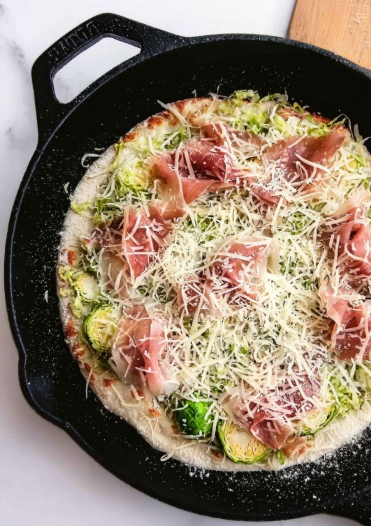 Unbaked pizza topped with Brussels sprouts, shredded cheese, and slices of prosciutto in a cast iron pan.