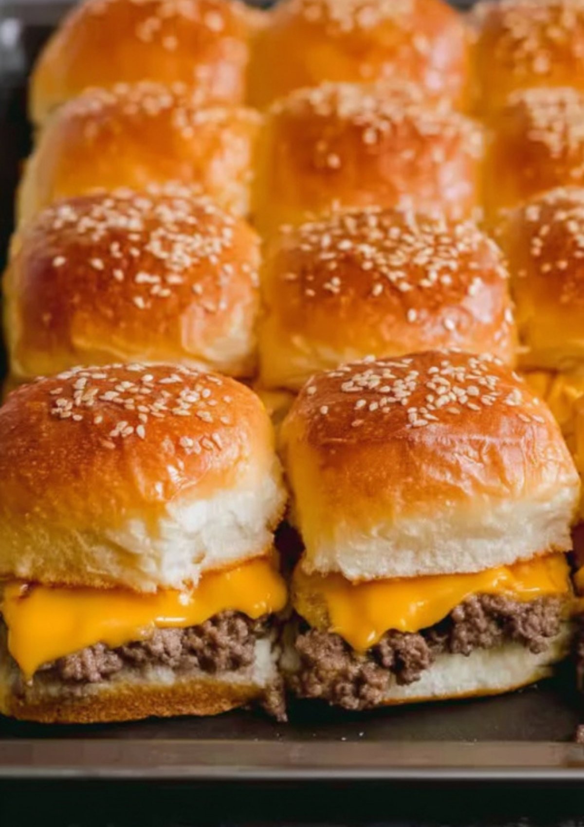Sheet pan of golden brown cheeseburger sliders with sesame seed tops, melted cheddar cheese, and juicy ground beef filling.