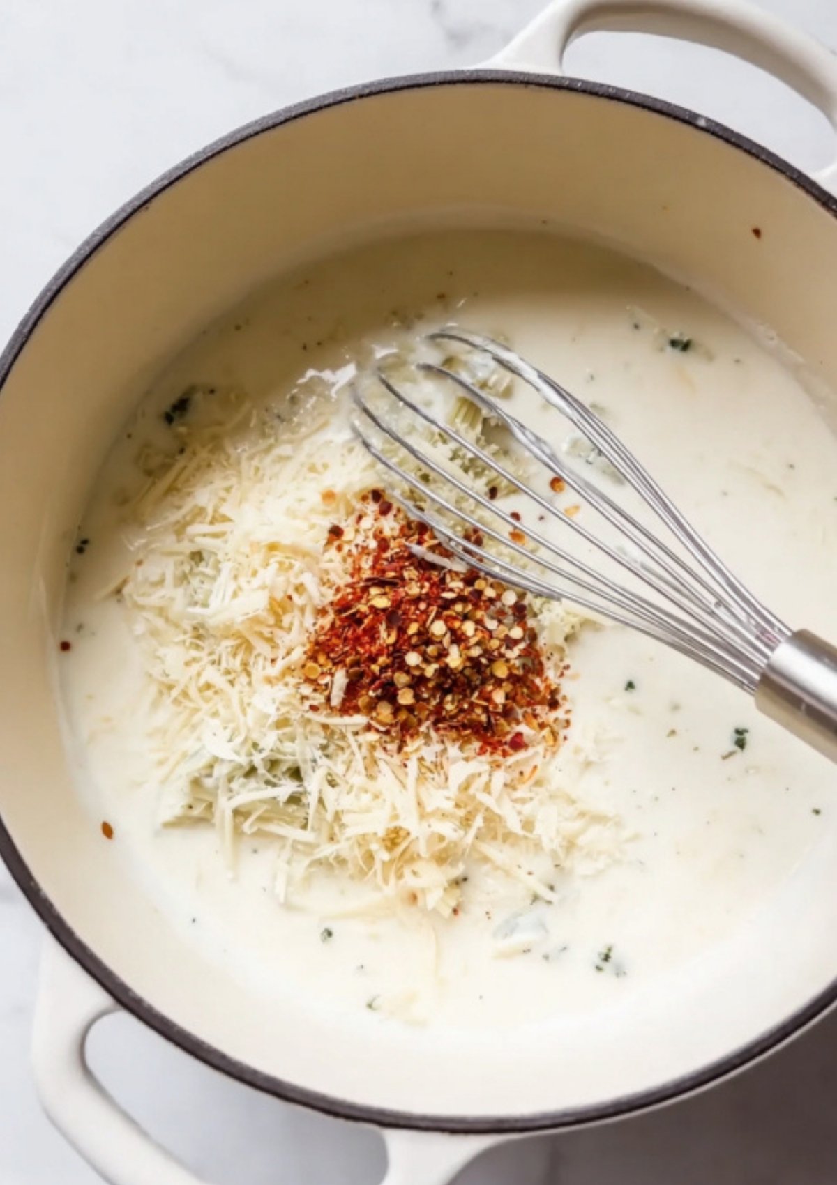 Dutch oven filled with cream sauce, shredded parmesan cheese, and red pepper flakes, ready to be stirred together.