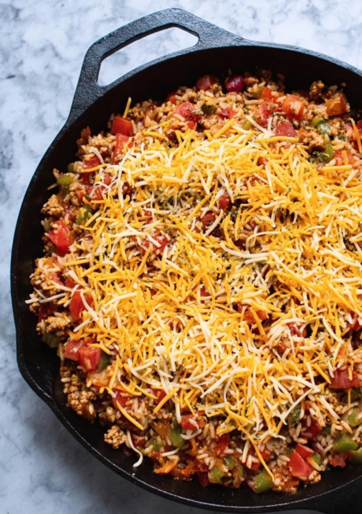 Skillet topped with shredded cheese and diced tomatoes, ready for baking or broiling until bubbly and golden.