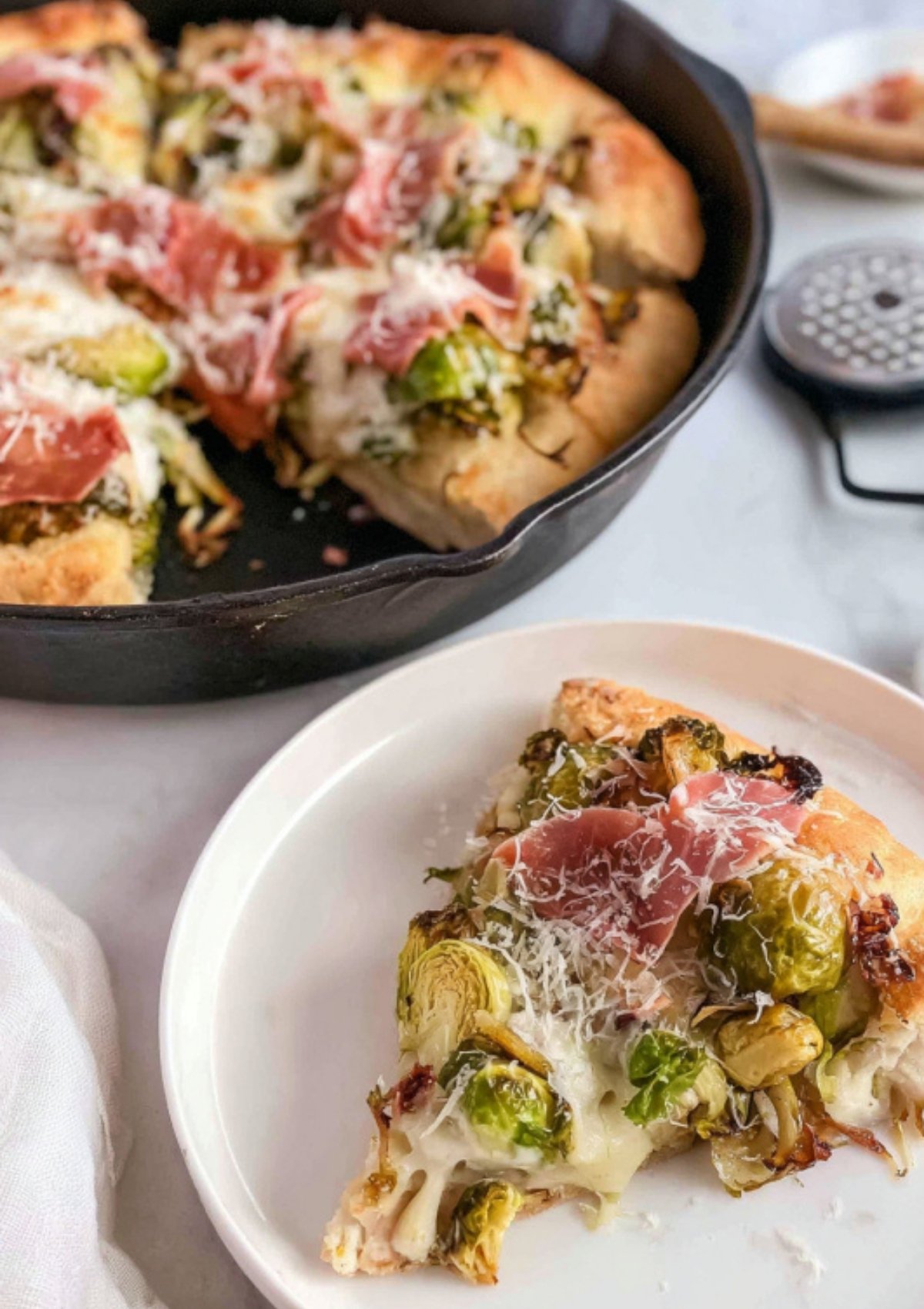 Slice of prosciutto pizza with roasted Brussels sprouts, melted cheese, and crispy crust served on a white plate beside the skillet pizza.