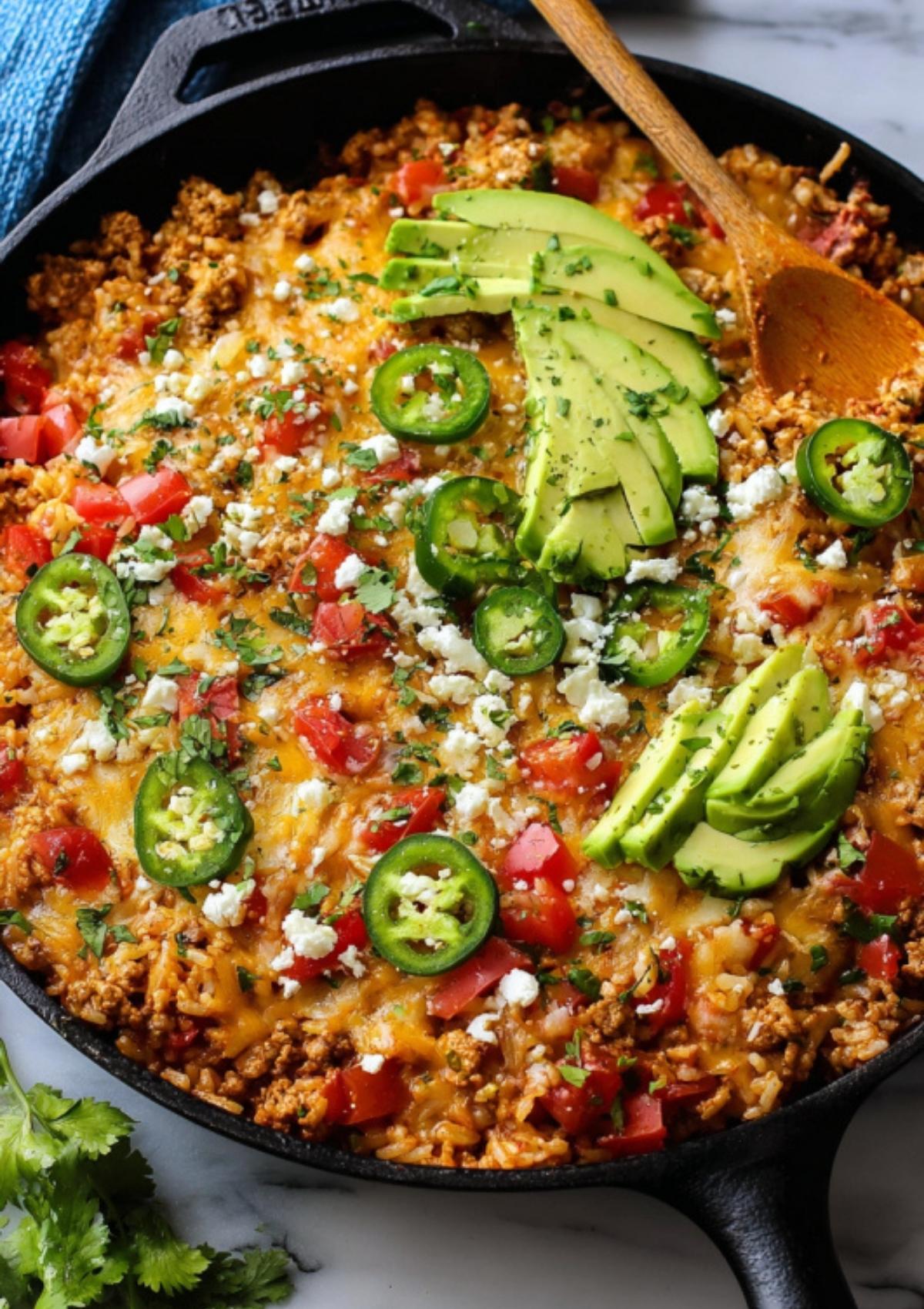 Hot skillet filled with cheesy ground turkey taco mix, garnished with cilantro, jalapeños, and sliced avocado.