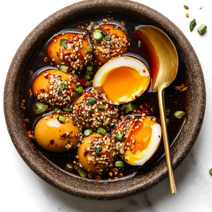 Bowl of Korean mayak eggs (soy sauce marinated eggs) with jammy yolks, topped with sesame seeds, green onions, and chili flakes, served with a gold spoon.