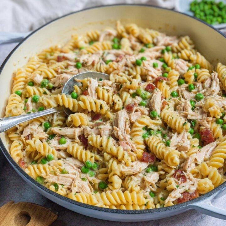 Creamy fusilli pasta with shredded roast chicken, peas, and bacon in a large white skillet with a serving spoon.