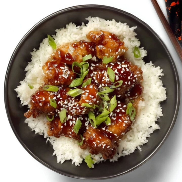 A black bowl of white rice topped with bite-sized pieces of soy glazed chicken, garnished with sliced green onions and sesame seeds.