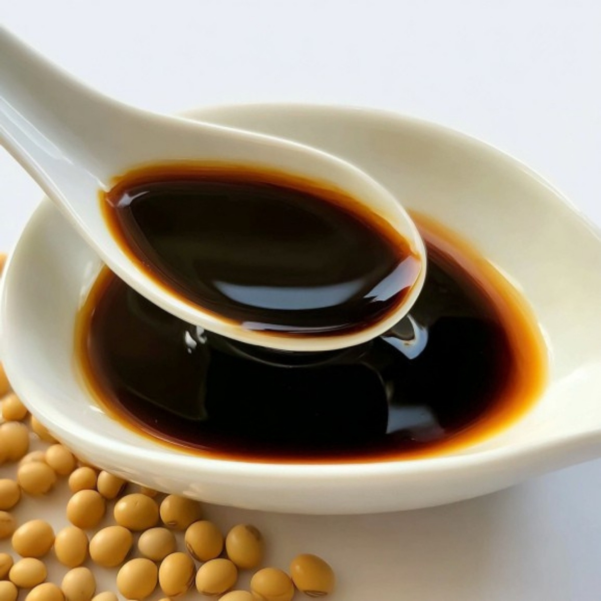 White ceramic spoon filled with rich, dark soya sauce resting in a matching bowl, with whole soybeans scattered nearby.
