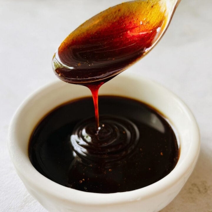 A close-up of a spoon swirling glossy, thick sweet soy sauce (kecap manis) in a white bowl, ideal for drizzling over fried rice or noodles.