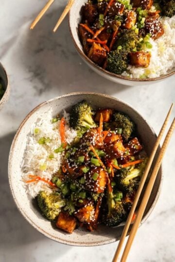 Two bowls filled with tofu stir-fry, broccoli, carrots, and white rice, topped with green onions and sesame seeds. Perfect for healthy vegan meal prep and plant-based dinner ideas.