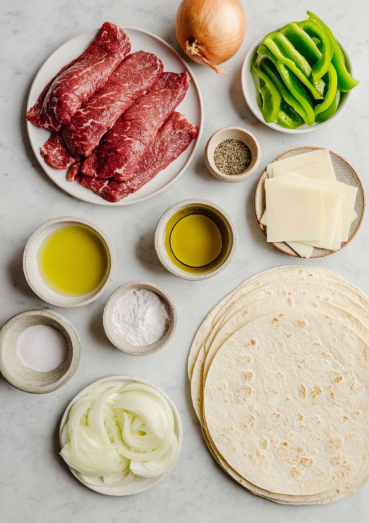Marble counter with steak, sliced peppers, onions, provolone cheese, tortillas, olive oil, and spices prepped for Philly cheesesteak wraps.