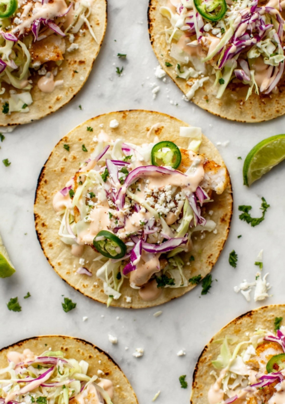 Street taco shells filled with crispy fish, cabbage, cheese, red onion, jalapeño, and a generous drizzle of creamy sauce, served with lime for taco night.