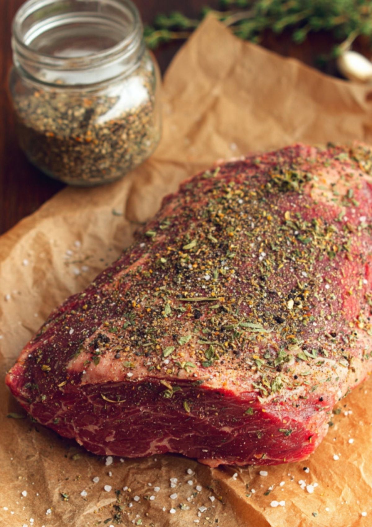 Raw chuck roast rubbed with spices and herbs on brown paper, jar of steak seasoning in the background