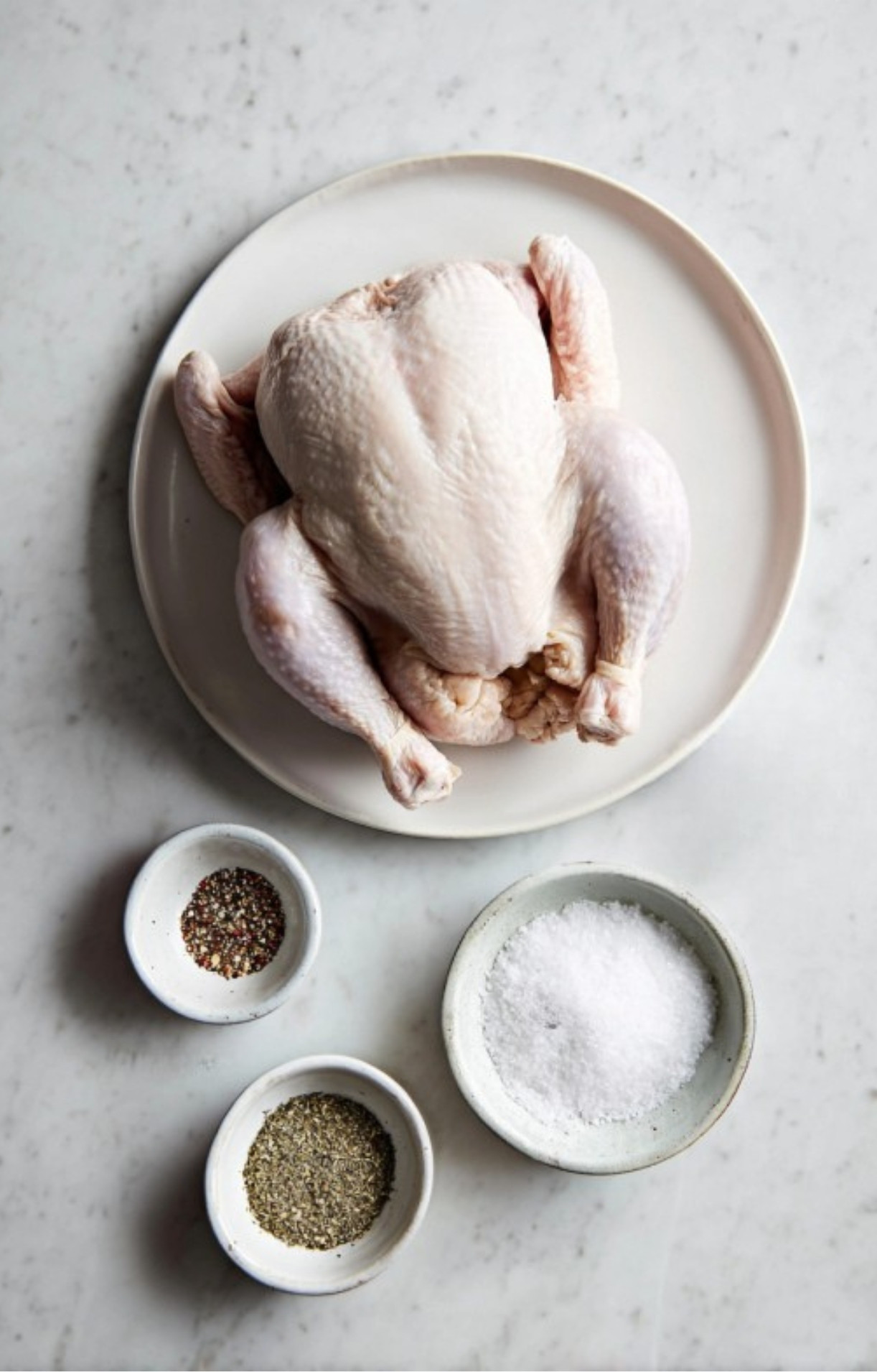 Uncooked whole chicken on white plate with bowls of salt, pepper, and Italian seasoning for classic roast chicken recipe