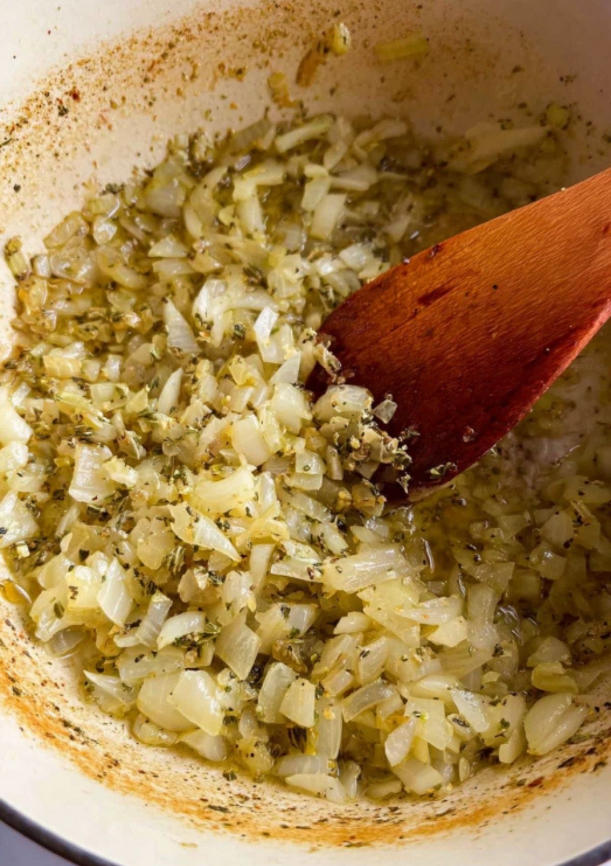Diced onions and Italian herbs sautéing in a pot with a wooden spoon for creamy tomato tortellini soup.