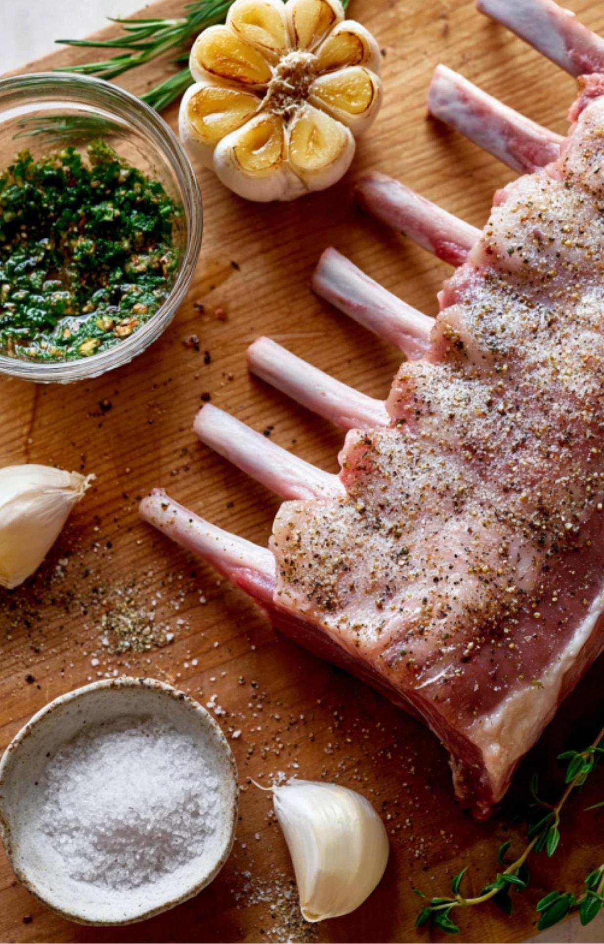 Uncooked rack of lamb seasoned with salt, pepper, and surrounded by roasted garlic and a bowl of herb paste.