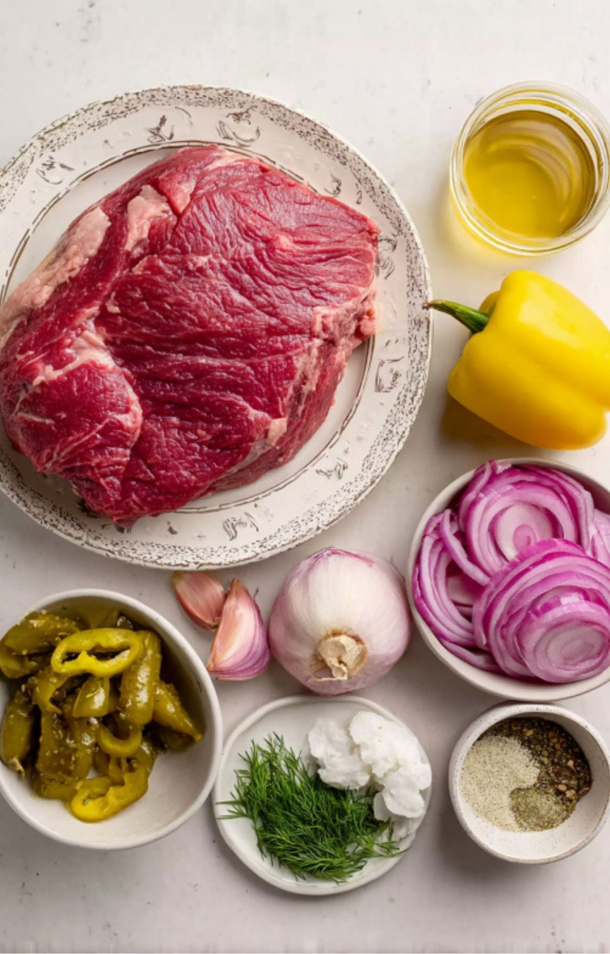 Overhead shot of Mississippi Pot Roast ingredients, including a beef chuck roast, yellow bell pepper, red onion, garlic, oil, fresh dill, pepperoncini, spices, and seasonings on a white background.