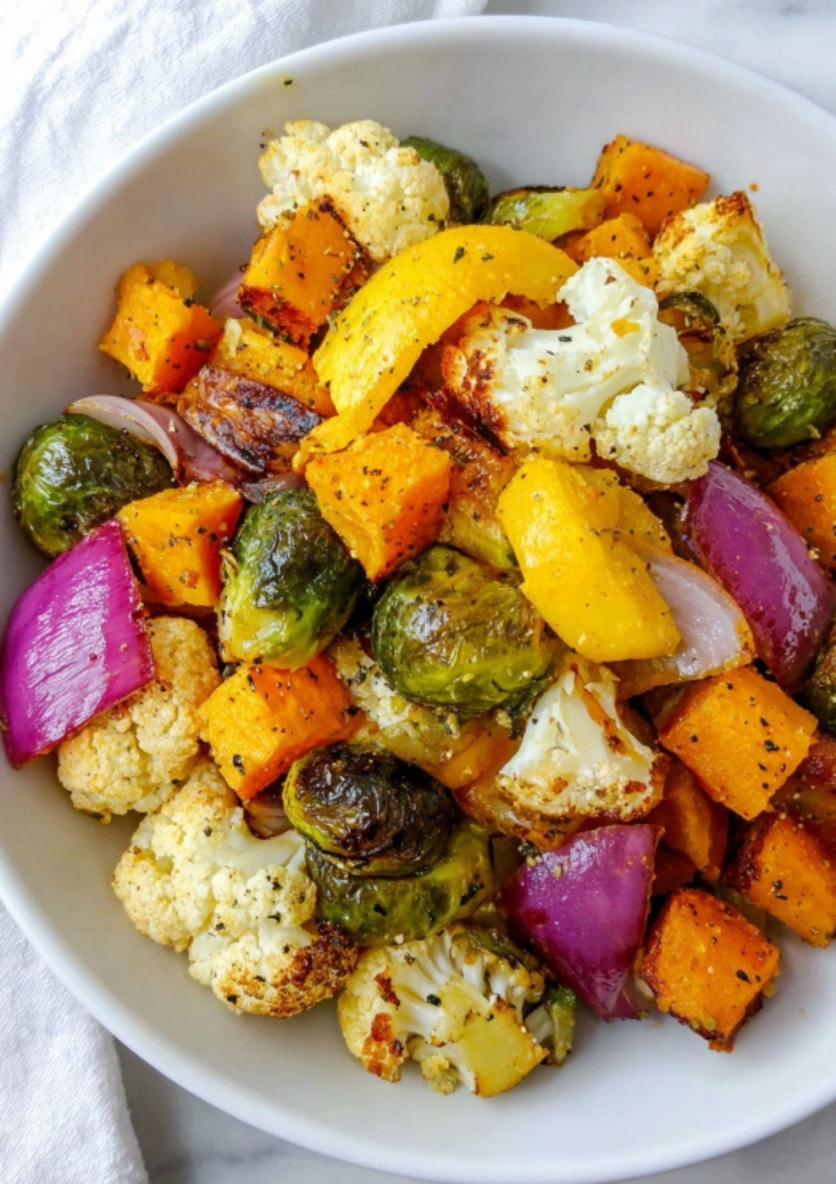 Closeup of a white bowl filled with oven-roasted red onion, cauliflower, yellow bell pepper, butternut squash, and brussels sprouts.