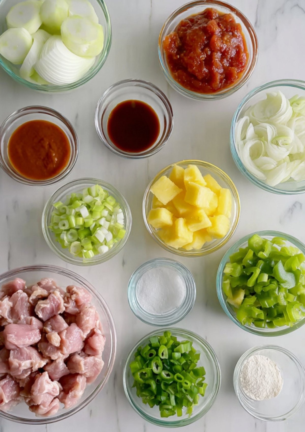 Flat lay of sweet and sour pork ingredients: pork cubes, pineapple, bell peppers, onions, green onions, sauces, and cornstarch.
