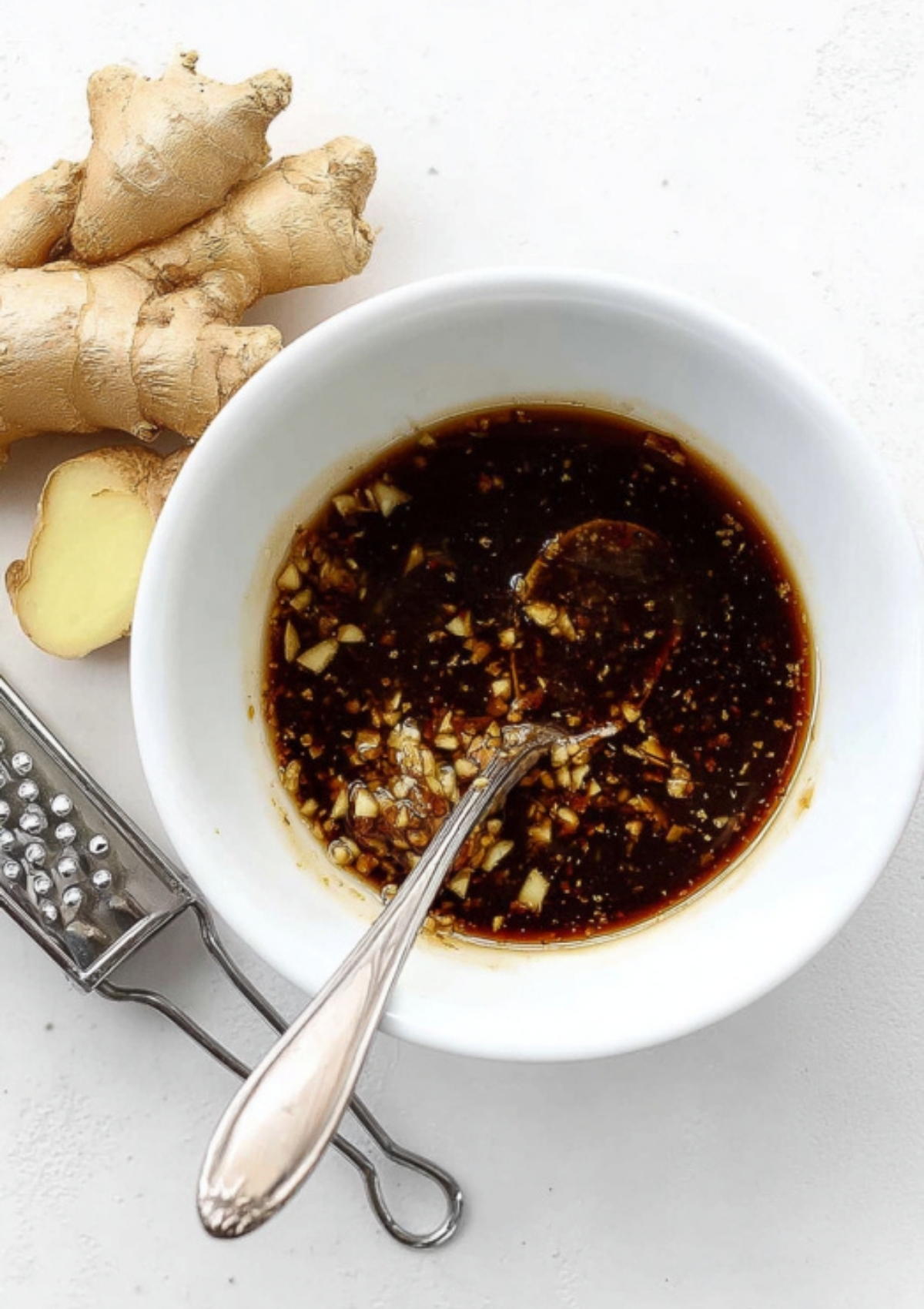 A small white bowl filled with a soy sauce-based marinade with minced ginger and garlic, next to a piece of fresh ginger and a grater.