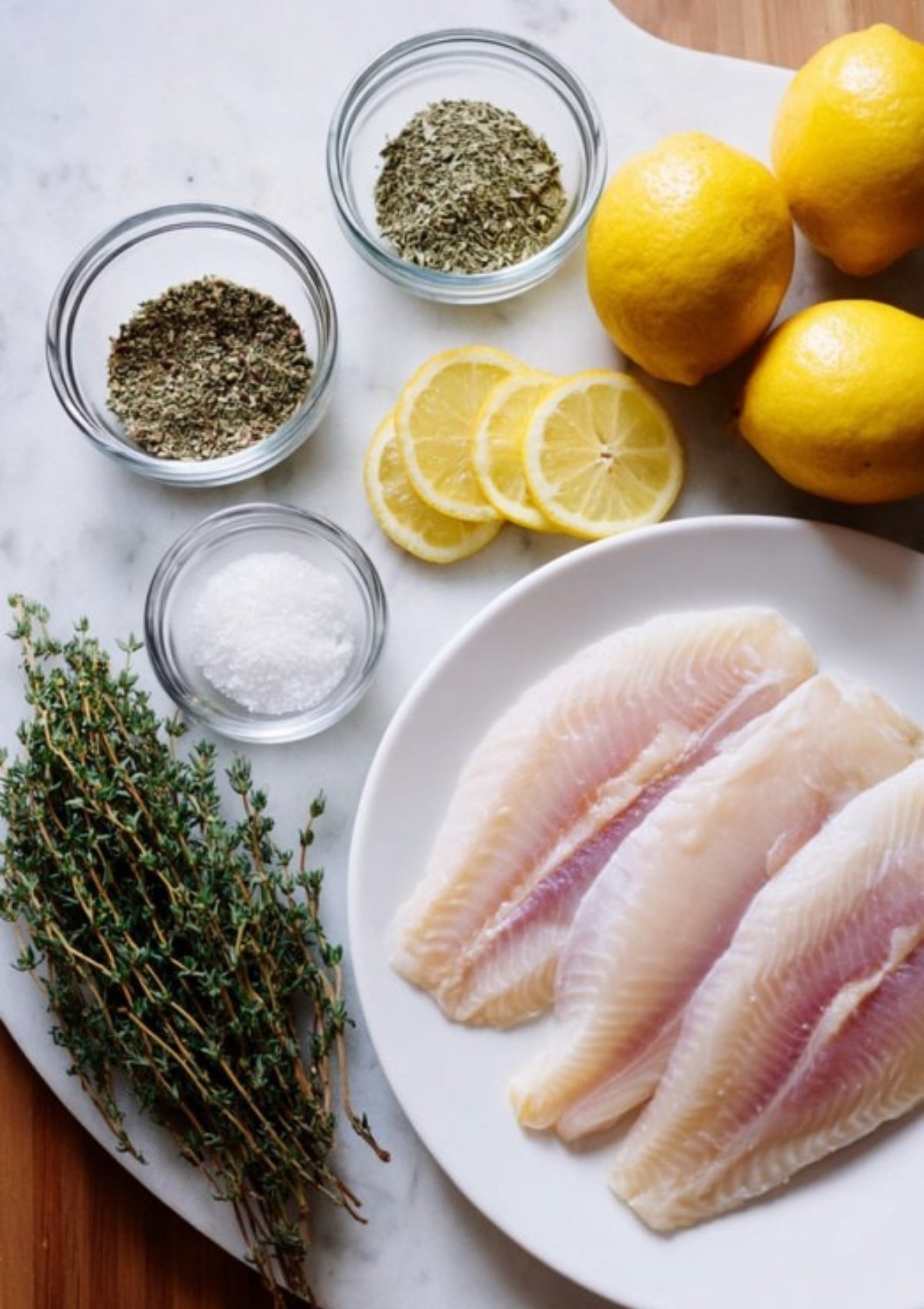 Marble board with tilapia fillets, lemons, fresh thyme, dried herbs, and salt—ingredients for lemon herb tilapia.
