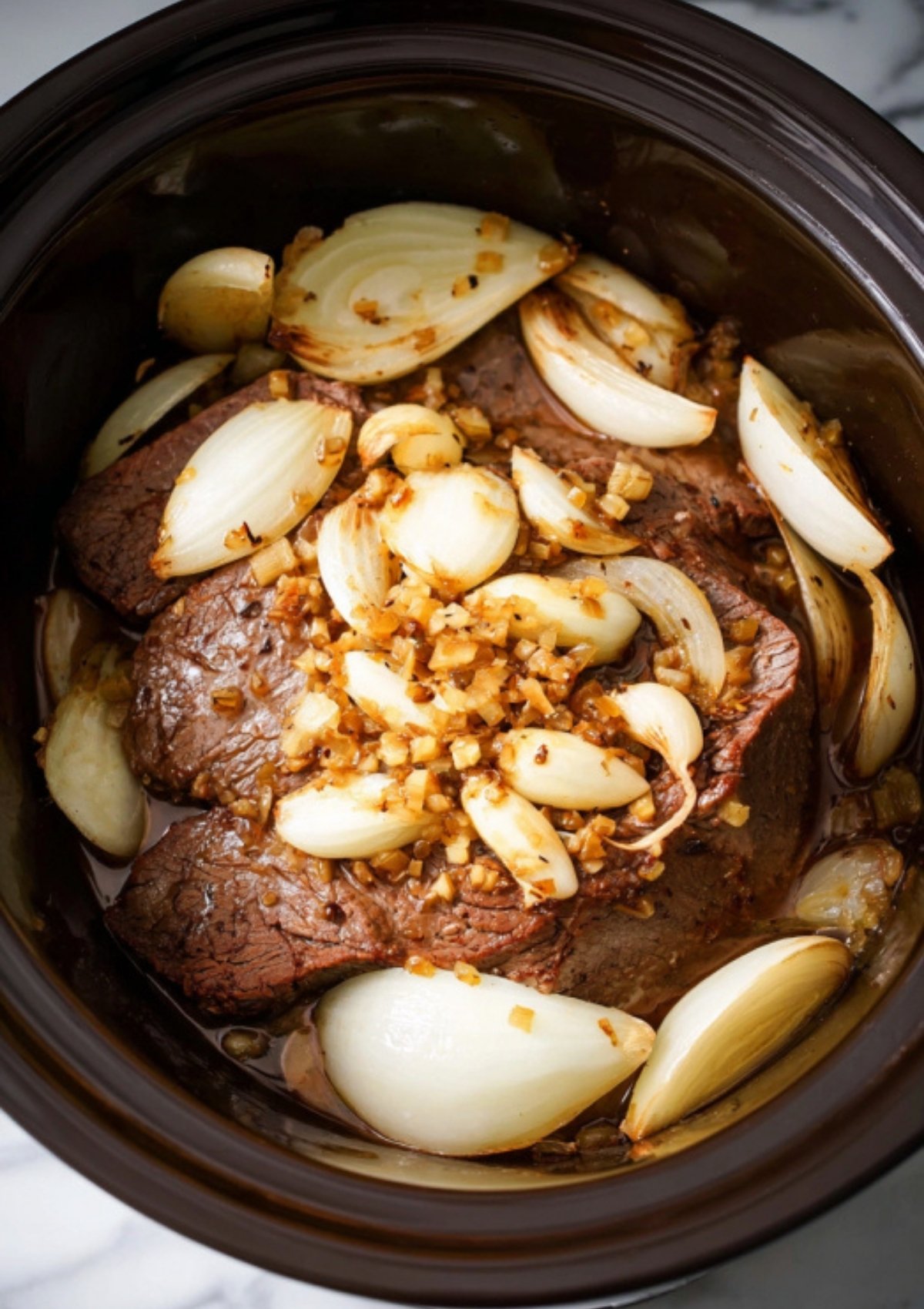 Beef roast in a crock pot topped with sliced onions and minced garlic, ready for slow cooking.