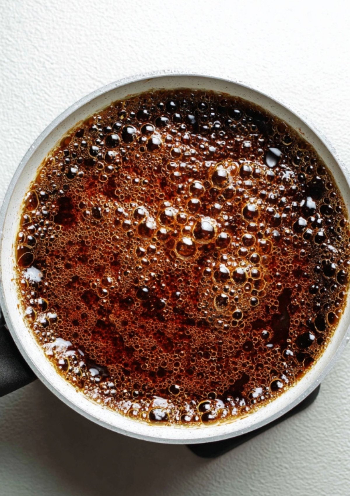 A small saucepan filled with bubbling, dark sweet soy sauce simmering on the stove, making homemade Indonesian kecap manis.