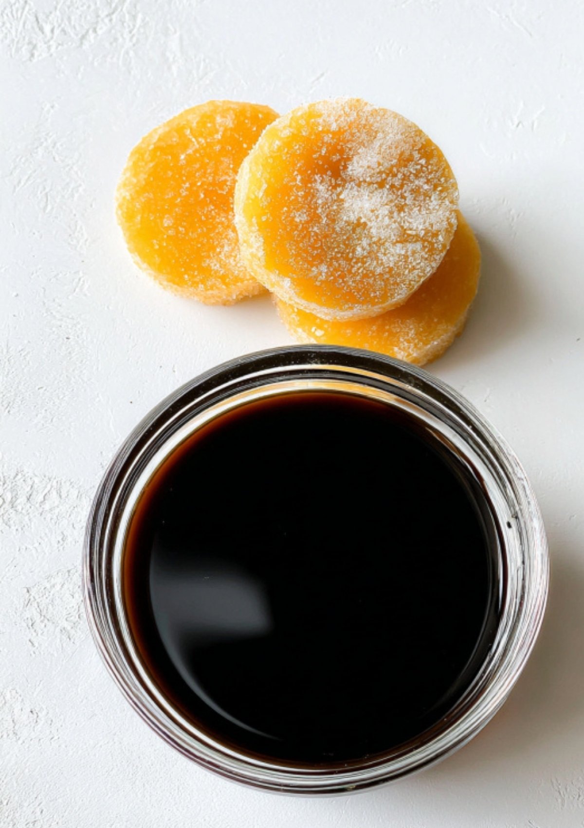 A bowl of dark sweet soy sauce next to rounds of palm sugar on a white background, showcasing the key ingredients for homemade kecap manis.