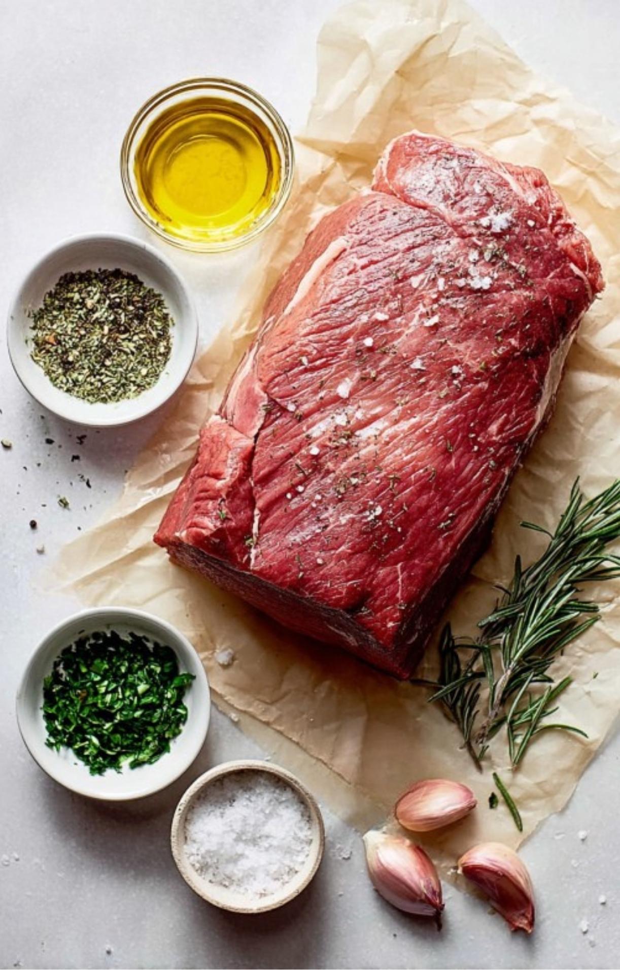 Raw beef roast on parchment surrounded by olive oil, fresh herbs, garlic, and salt