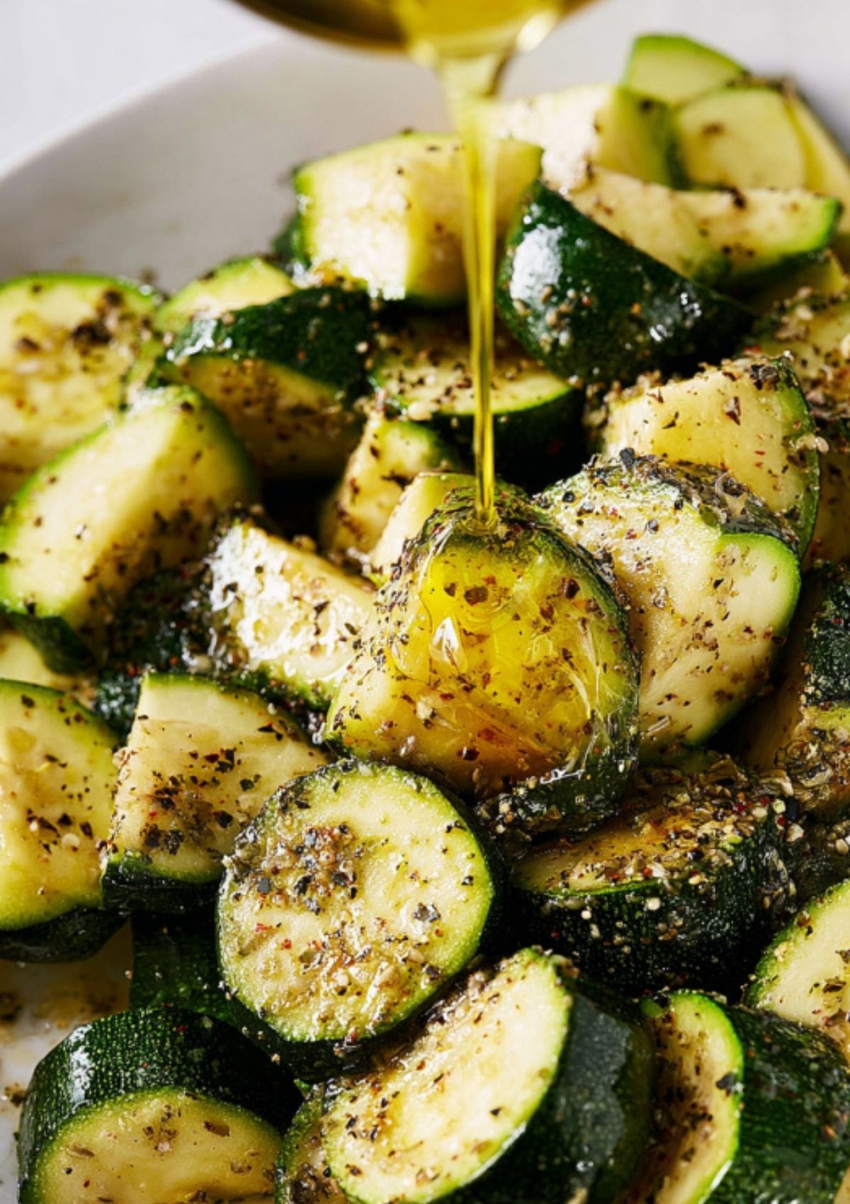 Closeup of olive oil being poured over seasoned zucchini slices in a white bowl.