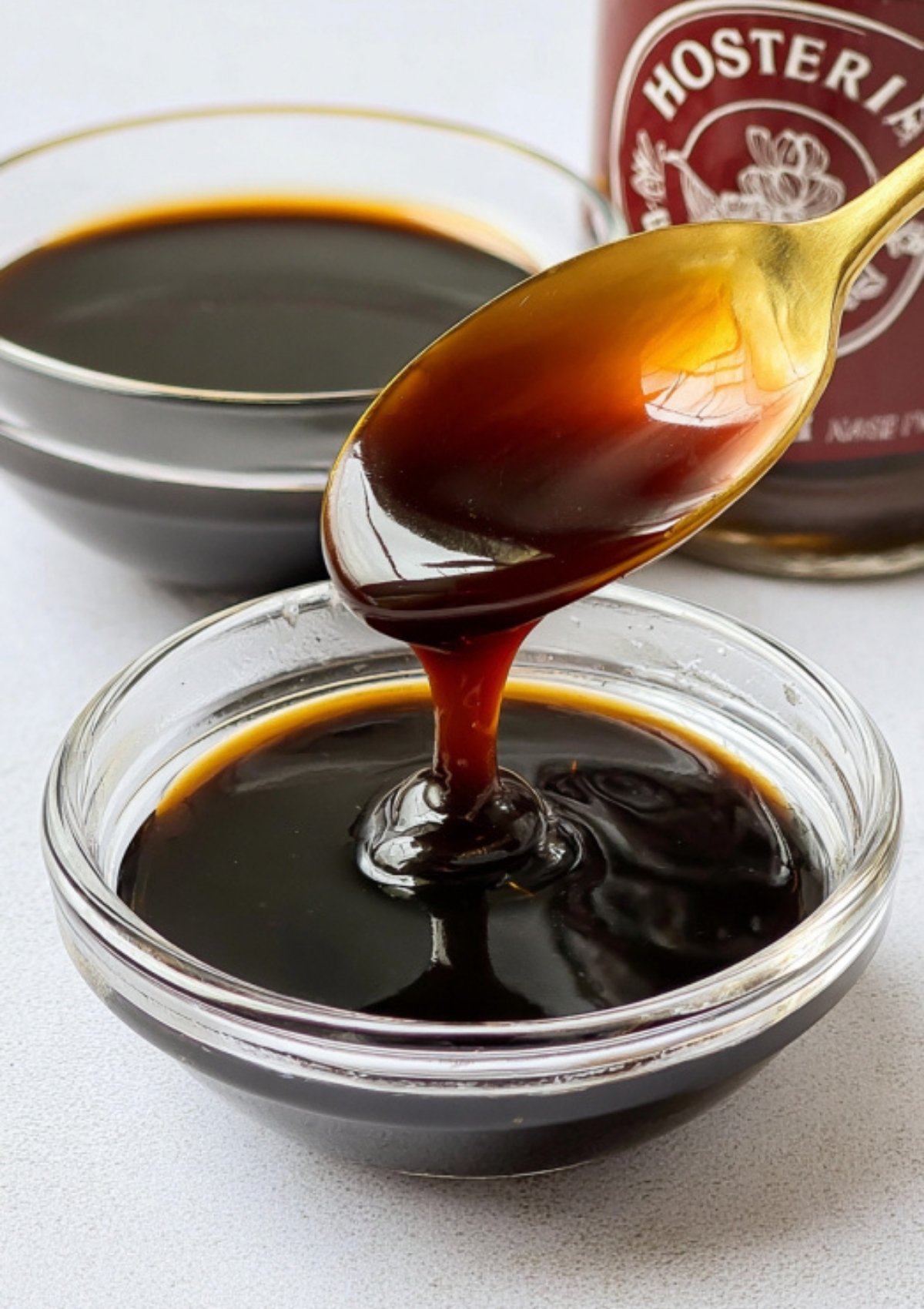 A gold spoon lifting thick, glossy homemade sweet soy sauce from a glass bowl, ready to drizzle over Asian dishes.