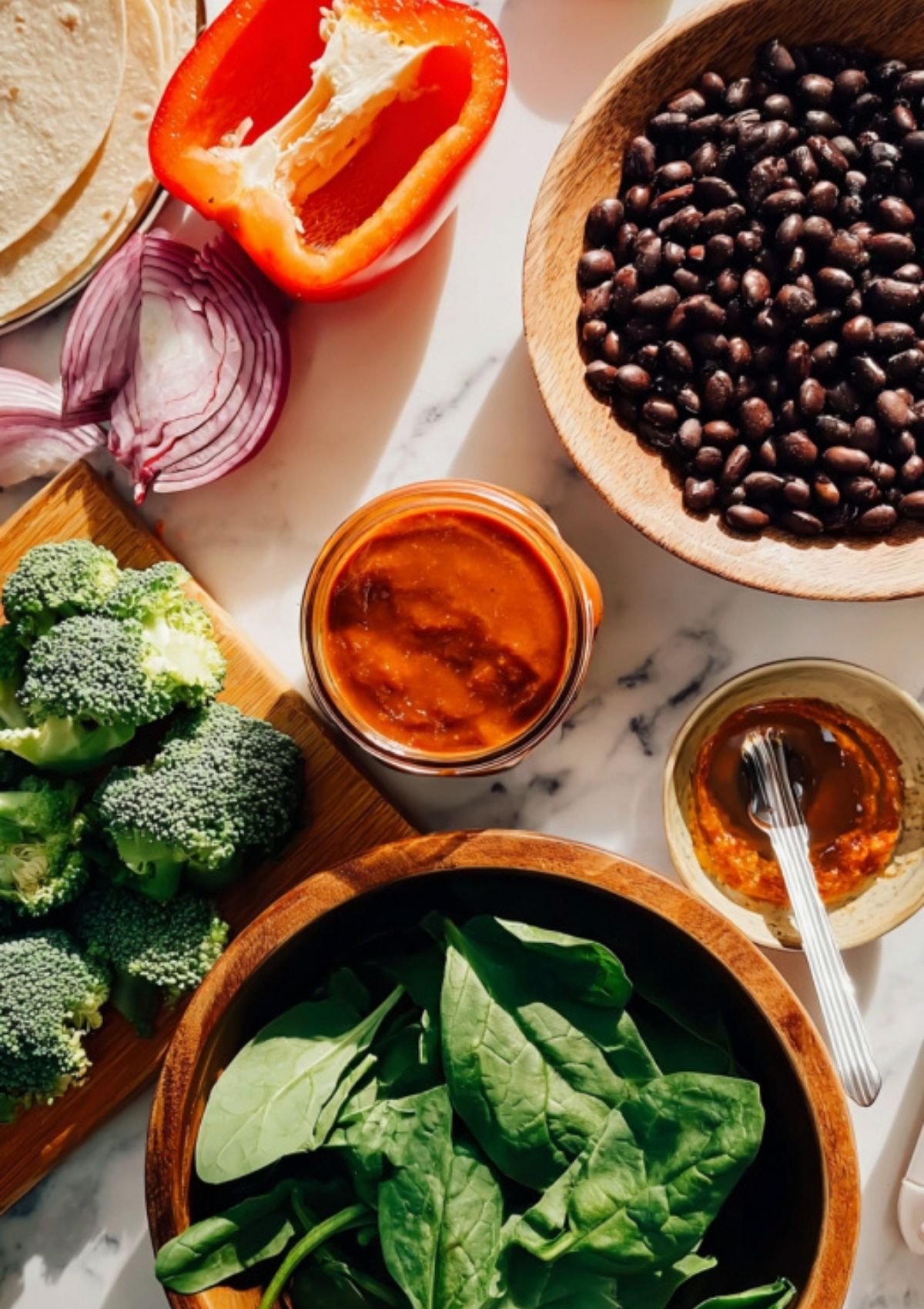 Wooden bowls with black beans, fresh spinach, broccoli, sliced red onion, red bell pepper, tortillas, and a jar of enchilada sauce—ingredients for black bean enchiladas.