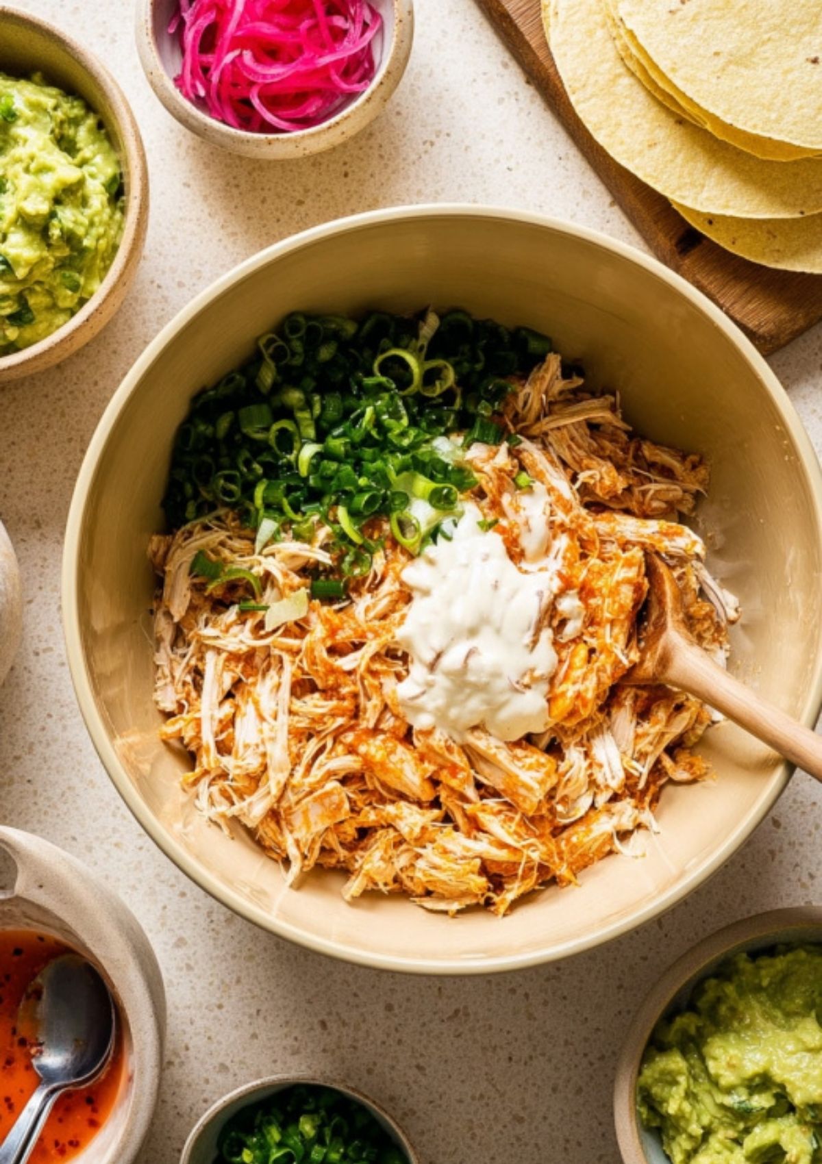 Bowl of shredded chicken mixed with buffalo sauce, green onions, and yogurt surrounded by guacamole, pickled onions, and tortillas.
