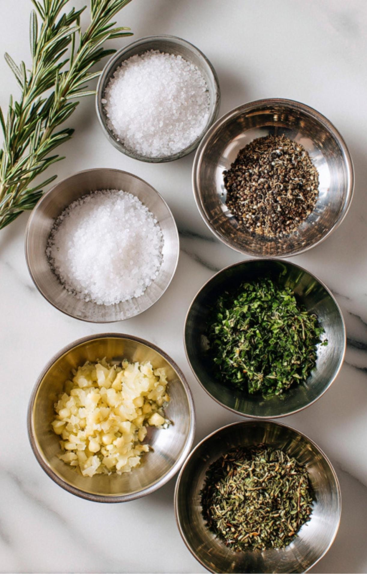 Bowls of kosher salt, cracked black pepper, garlic, rosemary, thyme, and parsley with fresh herbs for prime rib roast.