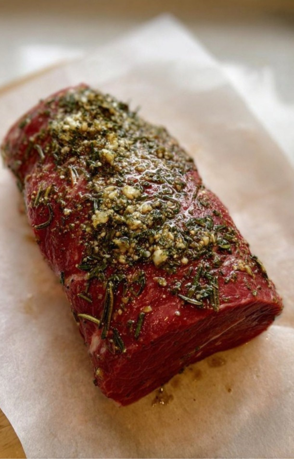 Uncooked beef roast on parchment, covered with fresh herbs, garlic, and olive oil marinade