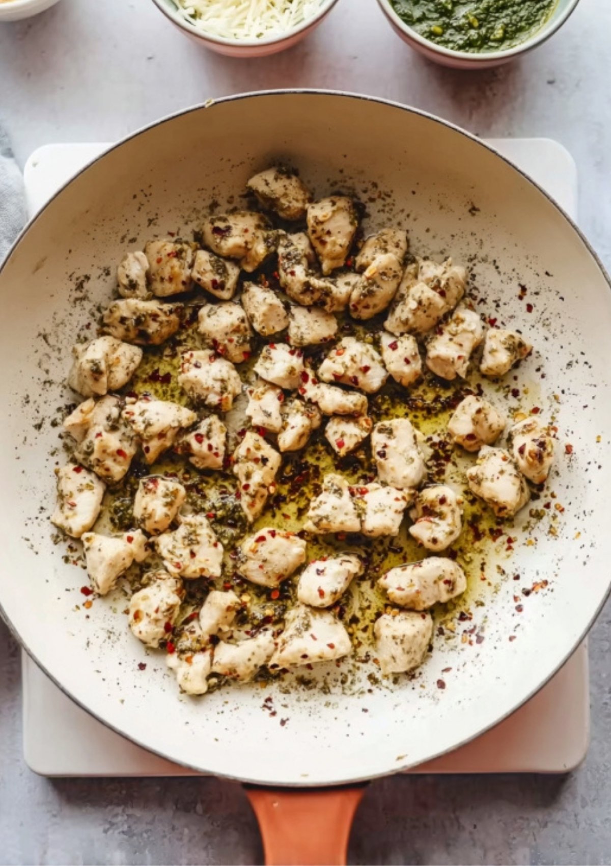 Seasoned chicken pieces cooking in a skillet for pesto chicken flatbread, surrounded by bowls of cheese, pesto, and tomatoes.